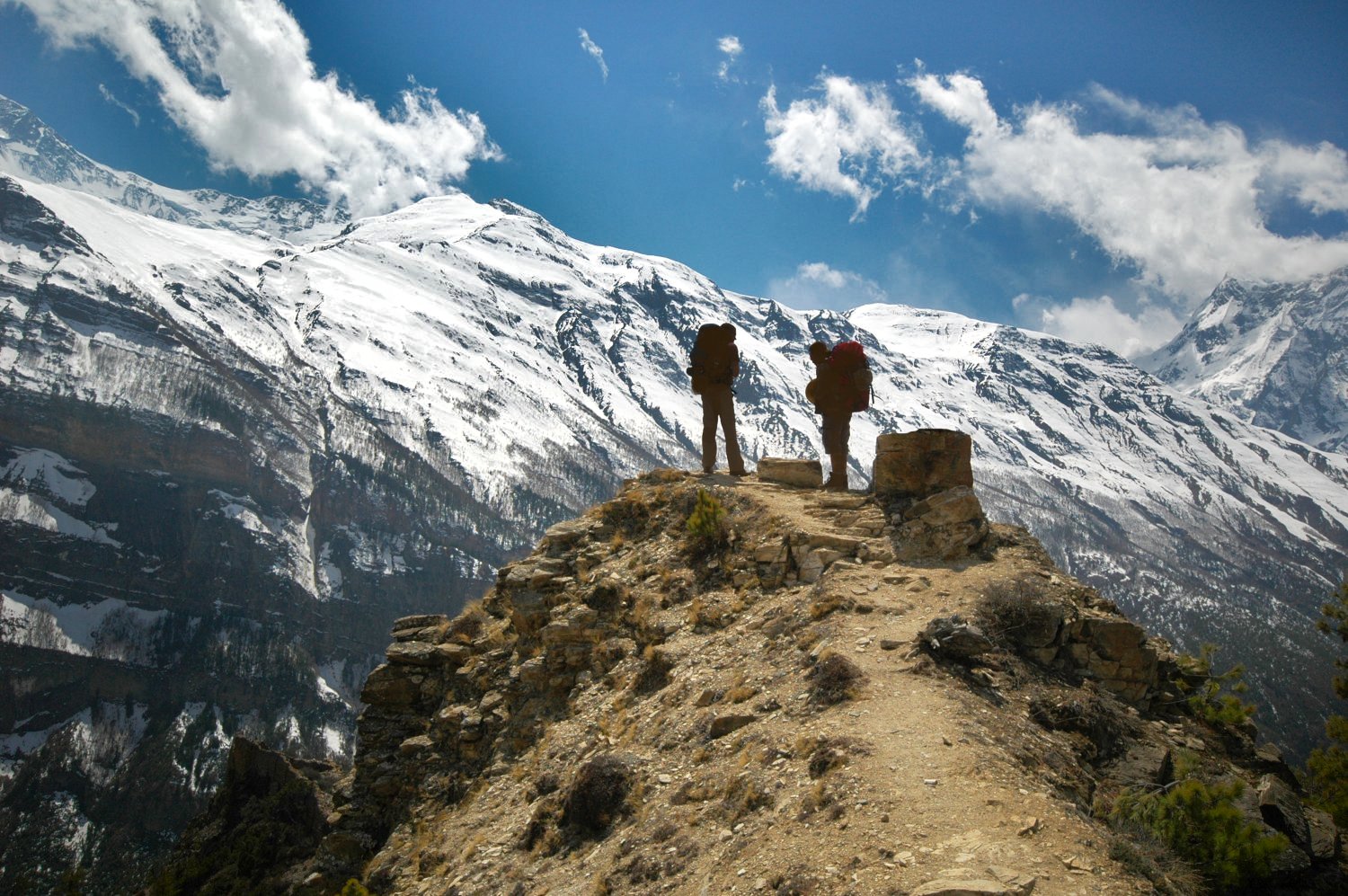 Two triumphant figures in hiking gear stand atop a peak with a snowy mountain range and a clear blue sky in the backdrop